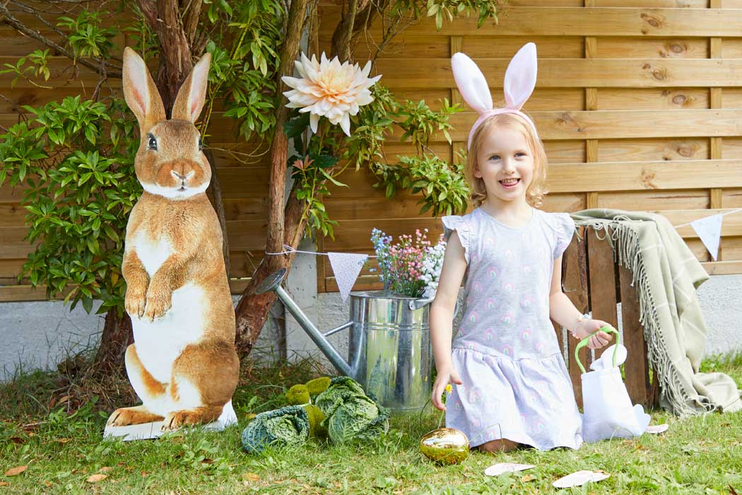 A young girl wearing bunny ears knelt on the grass in front of a fence, she is next to a cardboard cut out of Peter Rabbit amongst some othe