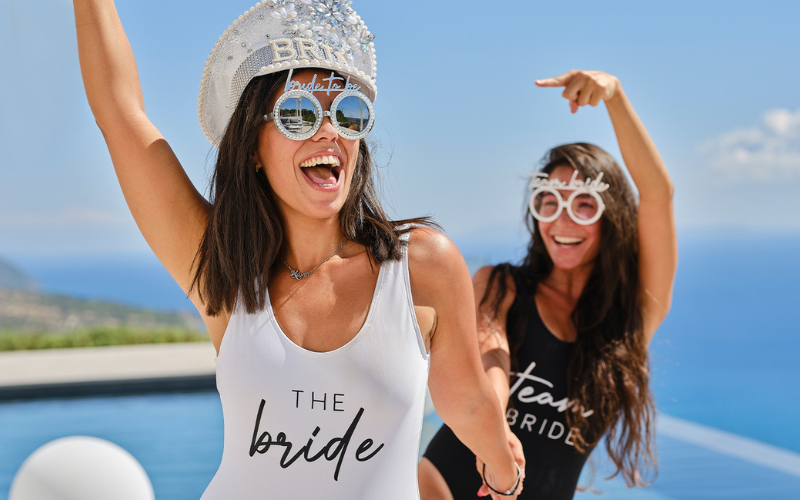 Bride dressed in white swimsuit with embellished hat dancing with bridesmaid in black swim suit and bride tribe sunglasses