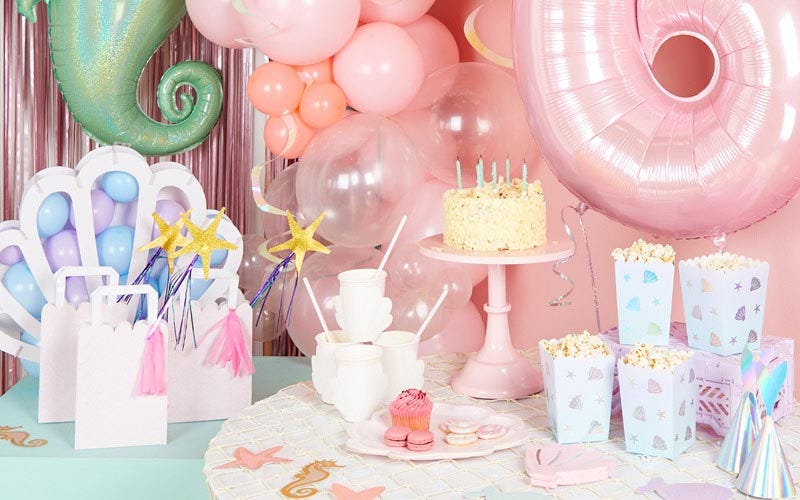 A mermaid birthday party setting with balloons, popcorn buckets, a cake and mermaid themed party bags