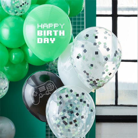 A bouquet of gaming balloons in front of a green background and a window on the right, there is a green balloon that reads 'happy birthday', come confetti balloons with black confetti inside and a black balloon with a gaming controller printed on it.