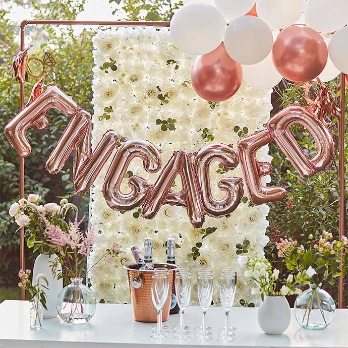 garden engagement party set up with champagne glasses and pink foil balloon banner spelling engaged