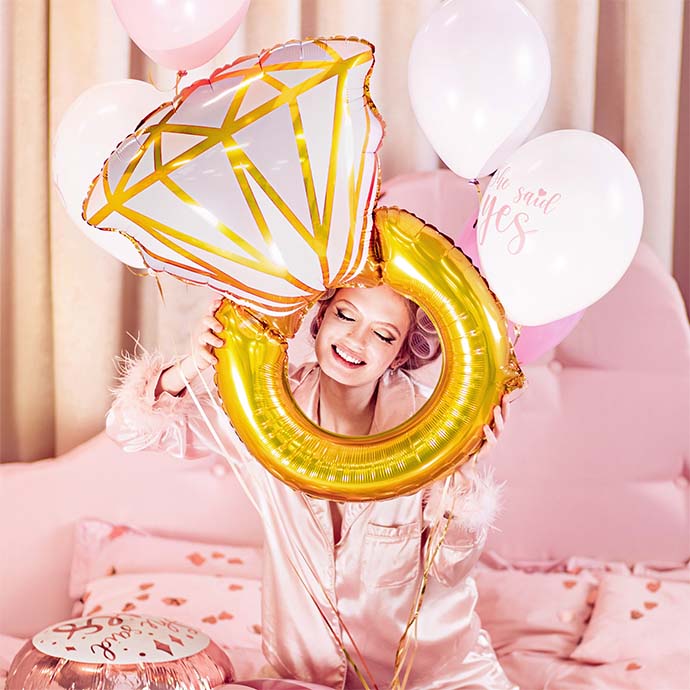 woman in pink pyjamas on a pink bed smiling with an engagement ring balloon