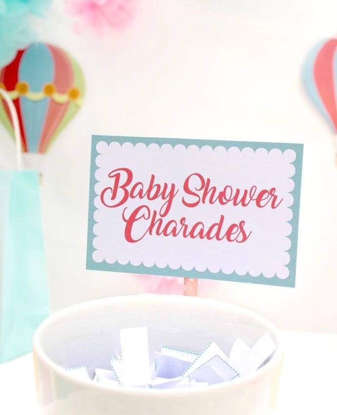 A bowl filled with folded up pieces of paper and a sign that says "Baby shower charades"