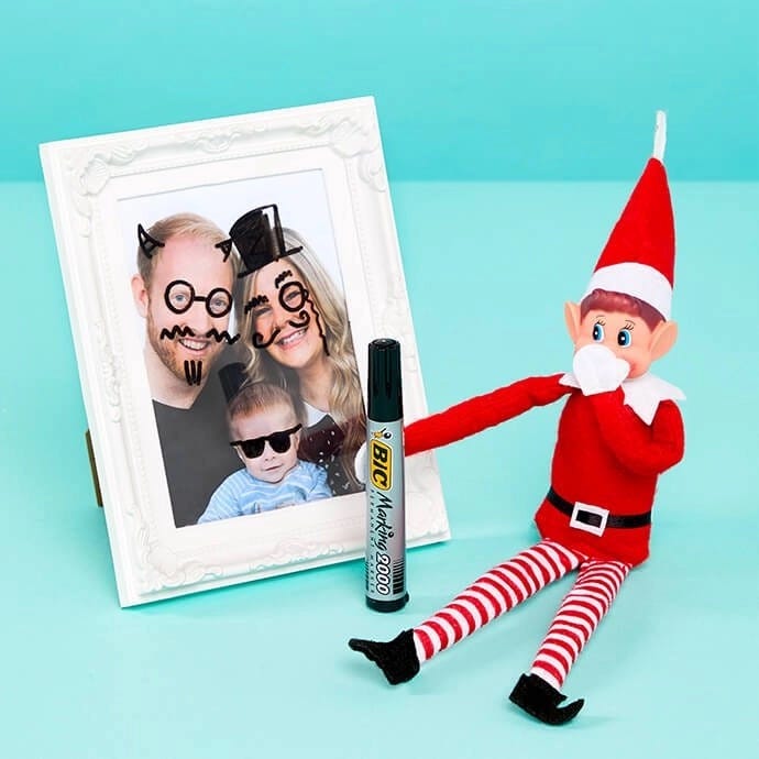 Elf on the Shelf drawing faces on a photo frame with a black marker pen